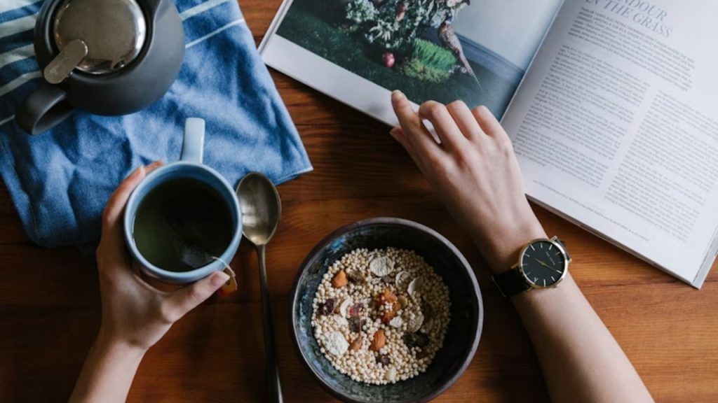 A person having tea and cereal while reading a magazine.