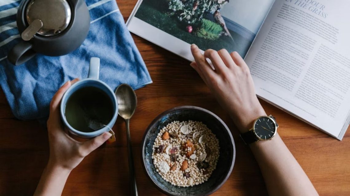 A person having tea and cereal while reading a magazine.