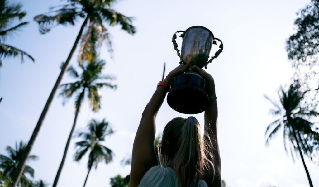 A person holding a trophy overhead among tall palm trees.