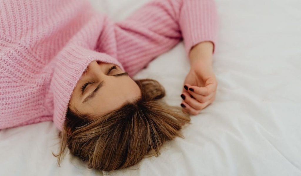 A person resting on a bed with a pink sweater covering part of their face.