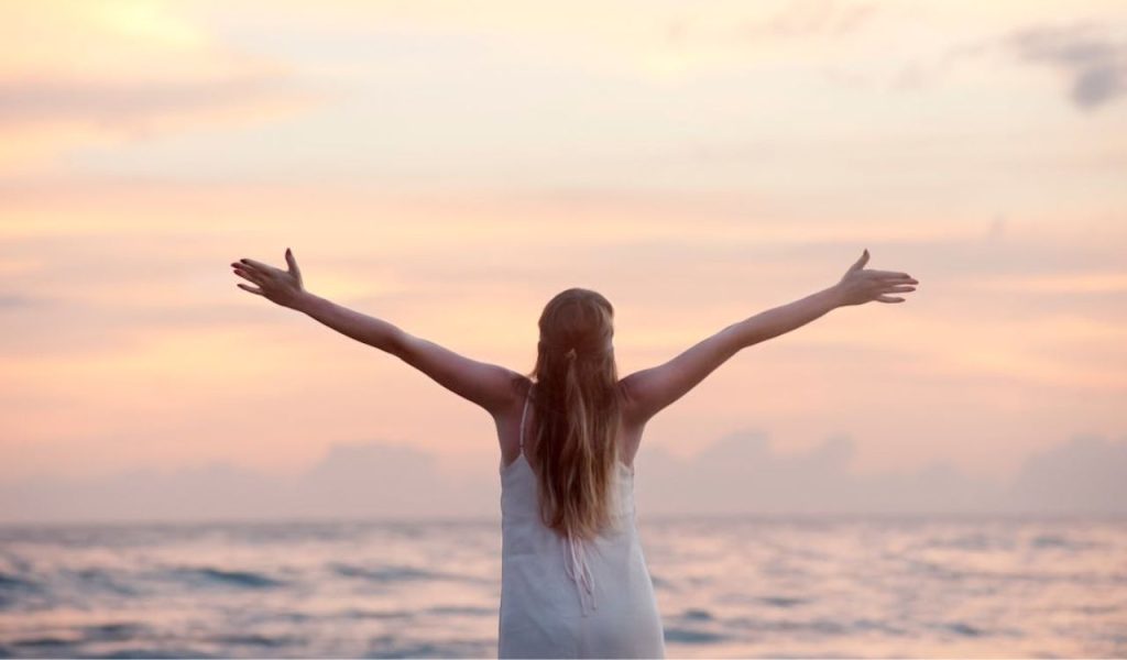 A person standing by the ocean with arms raised toward a pastel sunset.