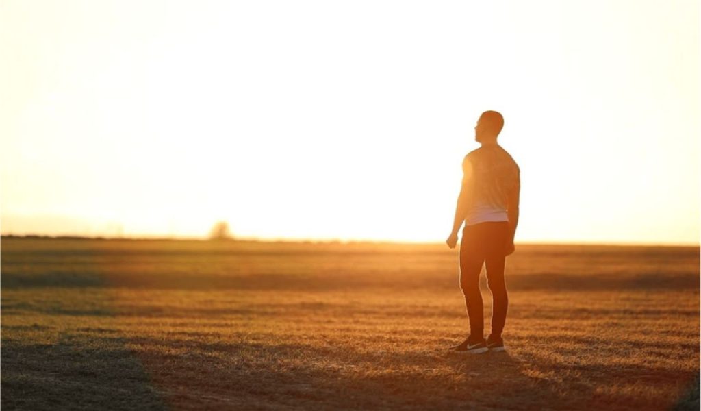 A person standing in a field at sunset, silhouetted by bright sunlight.