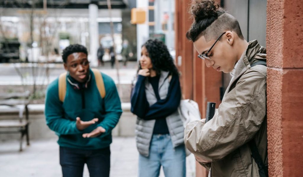 A person standing apart while two others talk and gesture nearby.