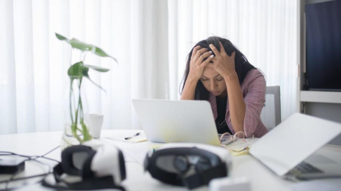 Overwhelmed woman at a desk, head in hands, staring at a laptop.