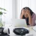 Overwhelmed woman at a desk, head in hands, staring at a laptop.