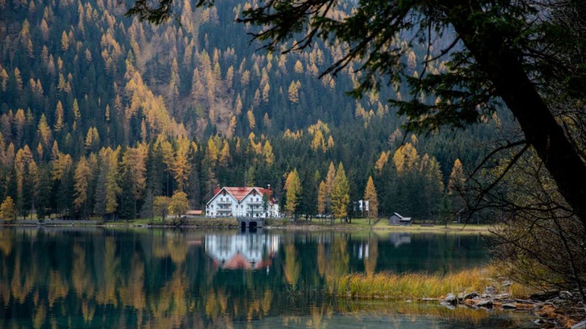 Quiet lake reflecting a white lodge with red roof, surrounded by dense pine forest and autumn trees.