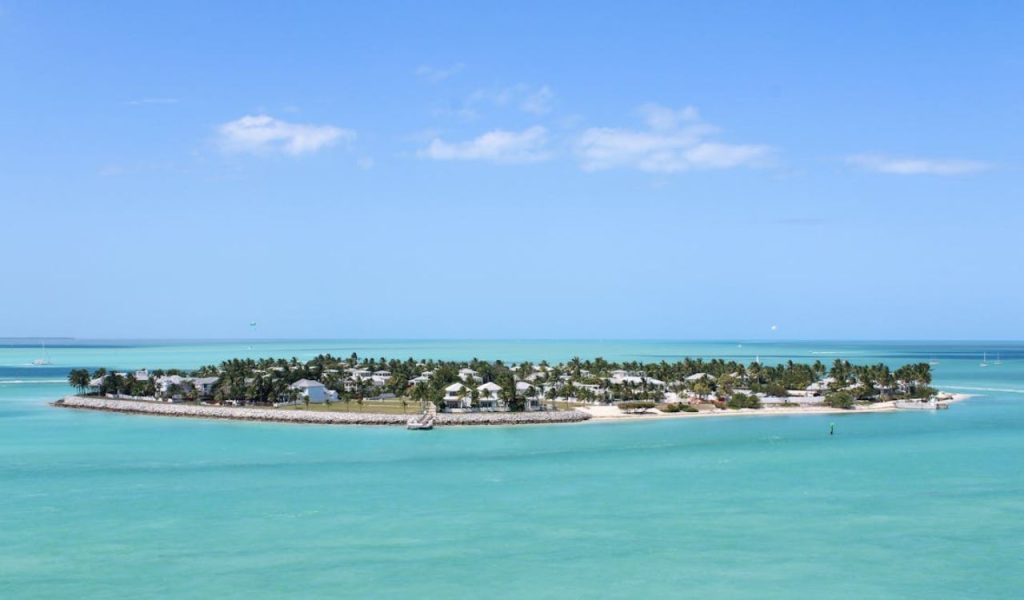 Small tropical island with palm trees in bright turquoise ocean under blue sky.