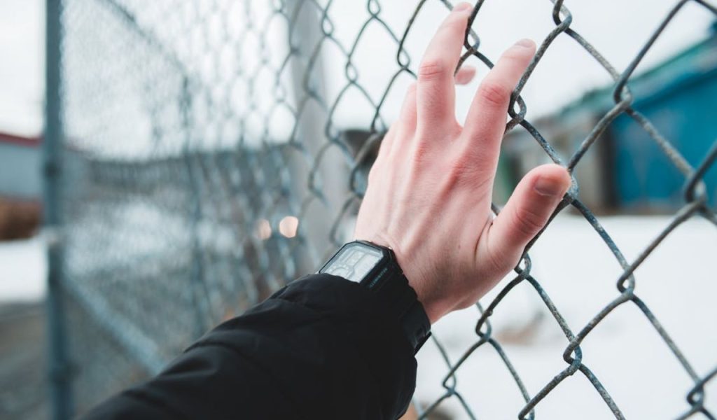 Close-up of a hand wearing a digital watch gripping a chain-link fence outdoors.