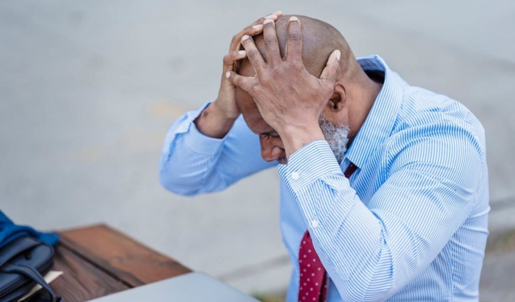 Stressed man sitting outdoors with hands on his head beside a laptop.