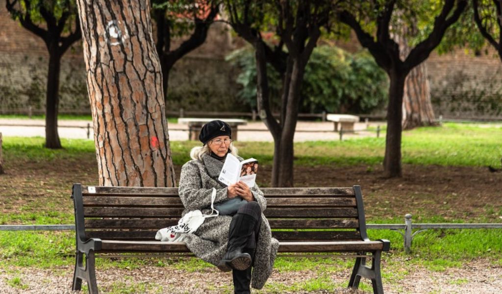 Elderly woman in a coat and beret reads a book while sitting on a wooden bench in a green park with trees.