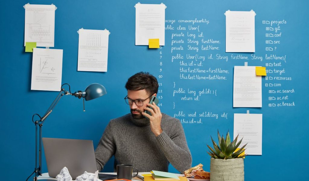 Man on the phone working at a laptop with papers taped to the wall.