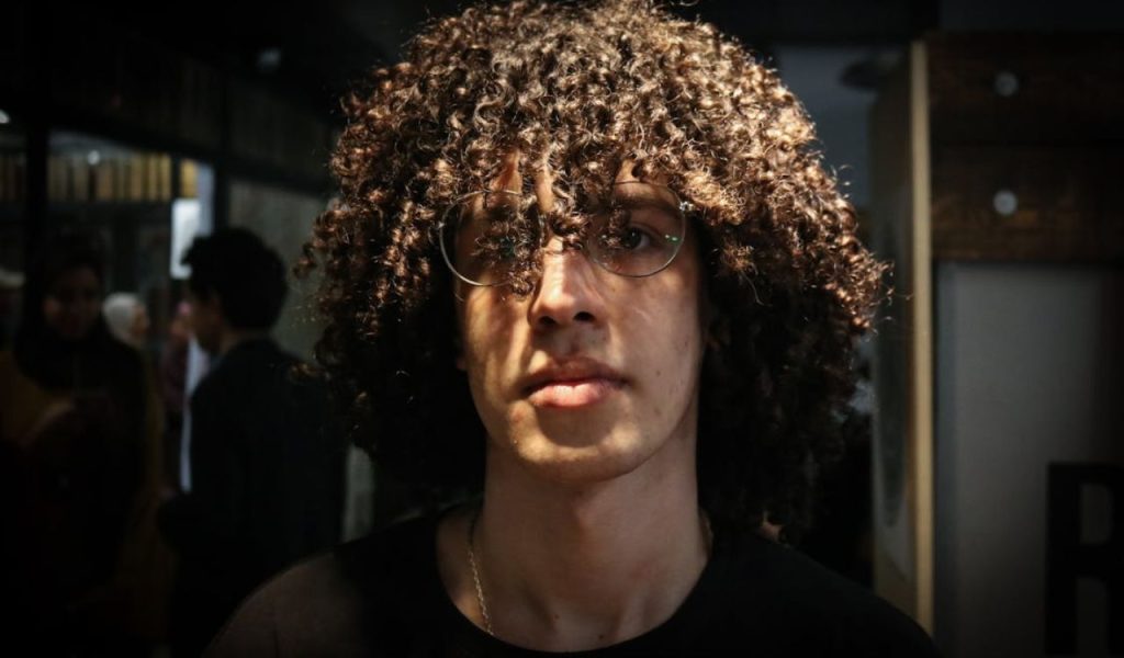 Curly-haired person wearing glasses, lit by strong sunlight, with a neutral expression.