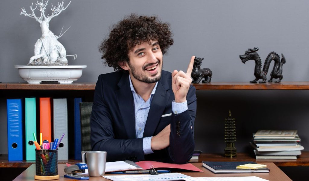 Smiling man at a desk pointing upward with one finger.