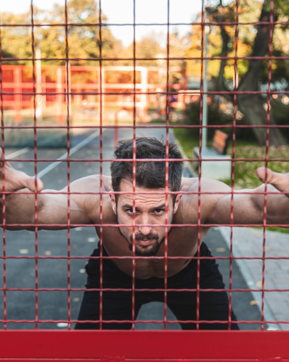 A muscular man pushes against a fence during an outdoor workout at a stadium.
