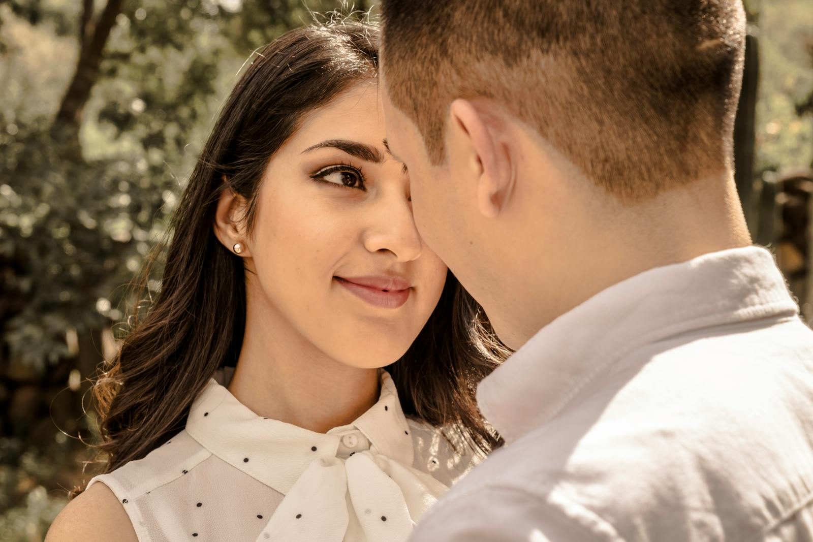 A young couple stands face to face, sharing a loving moment under a tree in daylight.