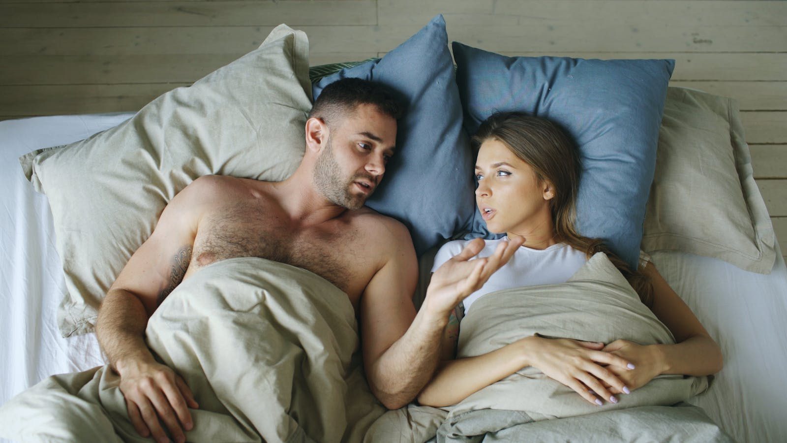 Couple lying in bed under blankets, engaged in conversation.