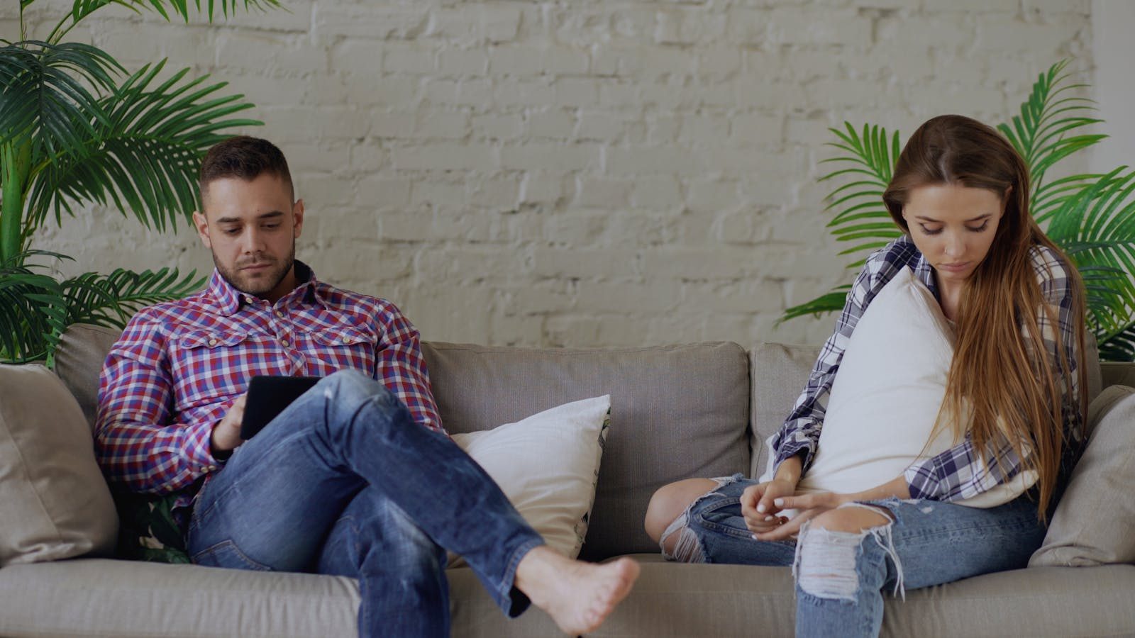A couple sits apart on a couch, engrossed in their thoughts or devices in a casual living room setting.