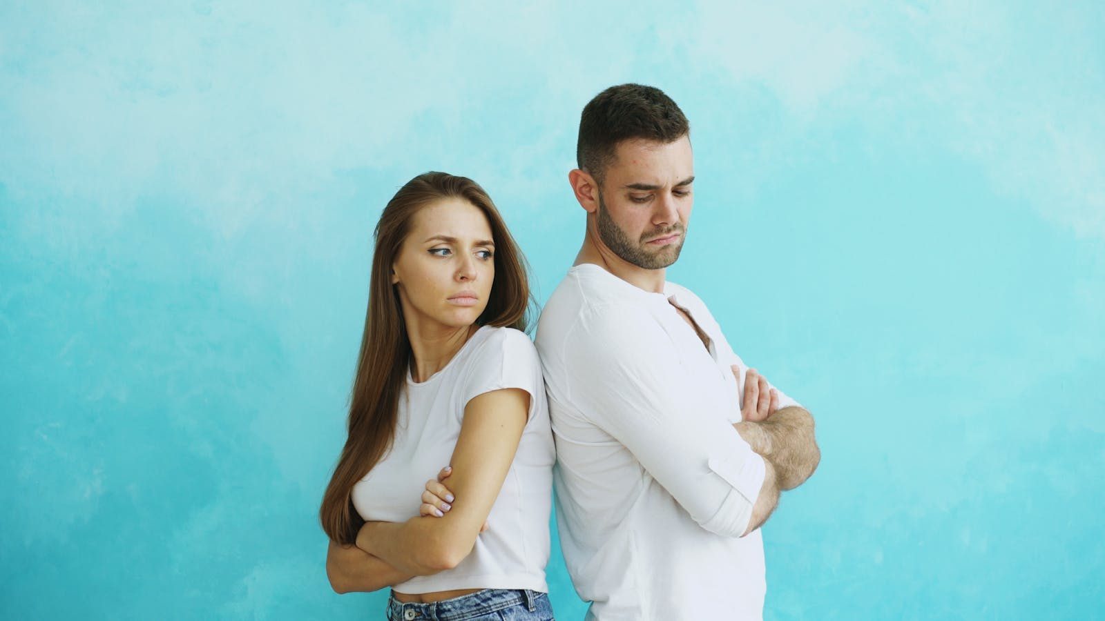 A tense couple stands back to back with crossed arms against a blue background.