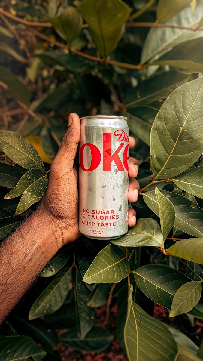 A hand holds a diet soda can amidst vibrant green leaves, creating a refreshing and natural contrast.