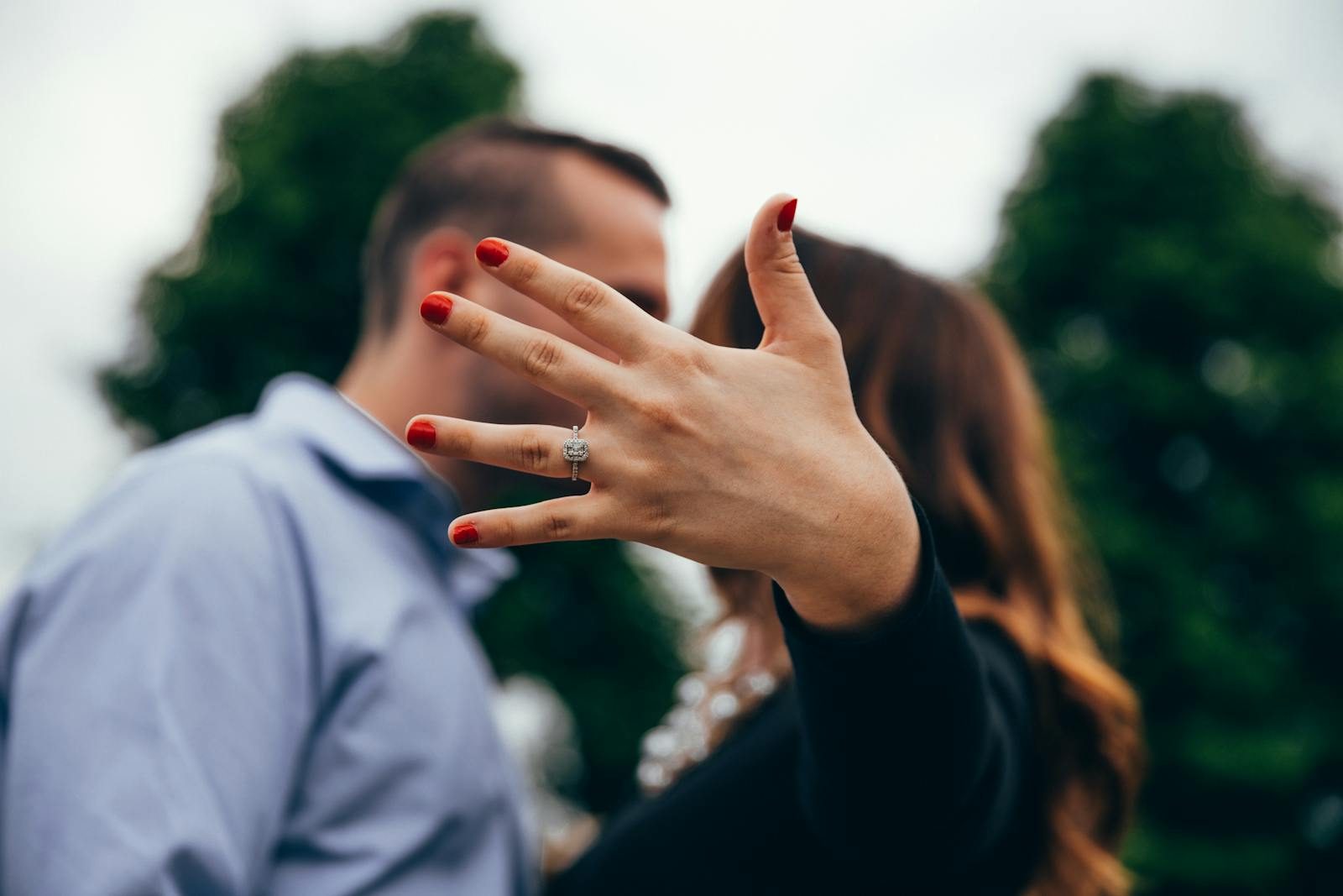 A close-up of a woman's hand showing off an engagement ring with a blurred couple in the background.