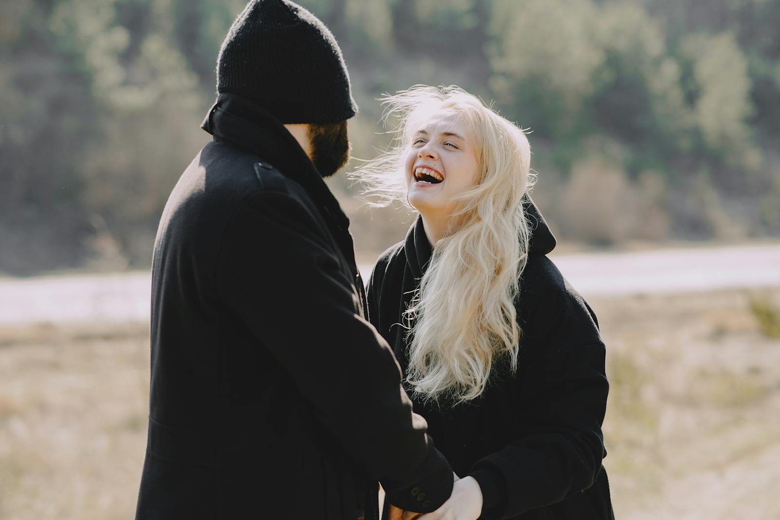Delighted laughing girlfriend in back lit in warm outerwear holding hands and smiling with boyfriend during stroll in sunny spring morning in countryside