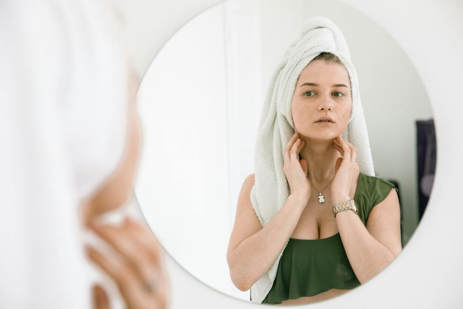 A woman with a head towel admires her reflection in a bathroom mirror, highlighting a beauty routine.