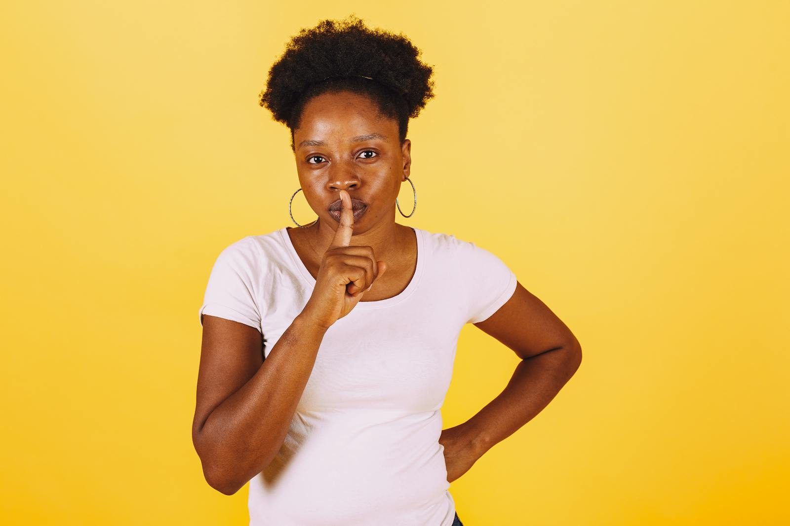 A woman in a white shirt making a silence gesture on a bright yellow background.