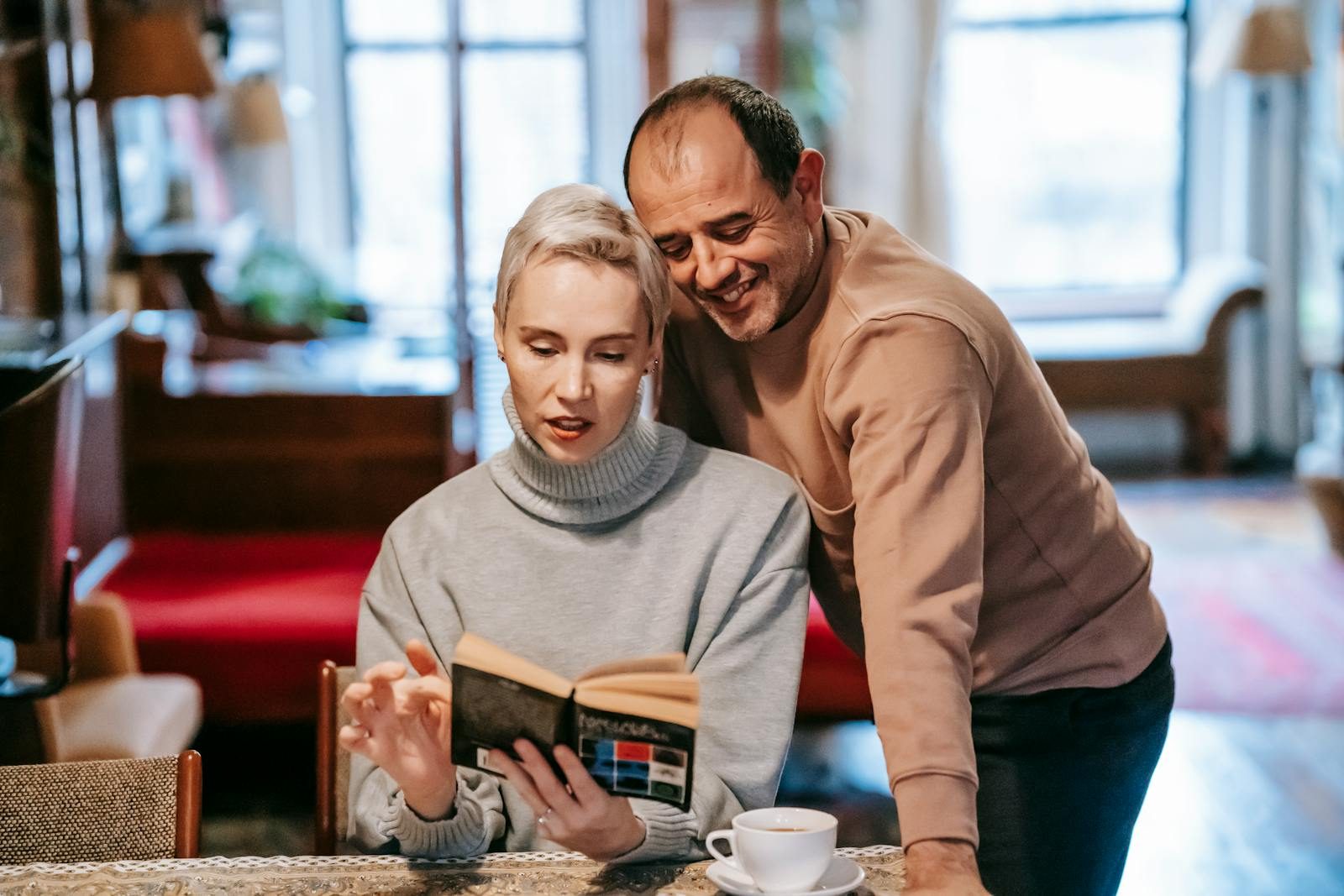Happy ethnic man leaning on wall near concentrated wife reading interesting book and drinking coffee during weekend at home