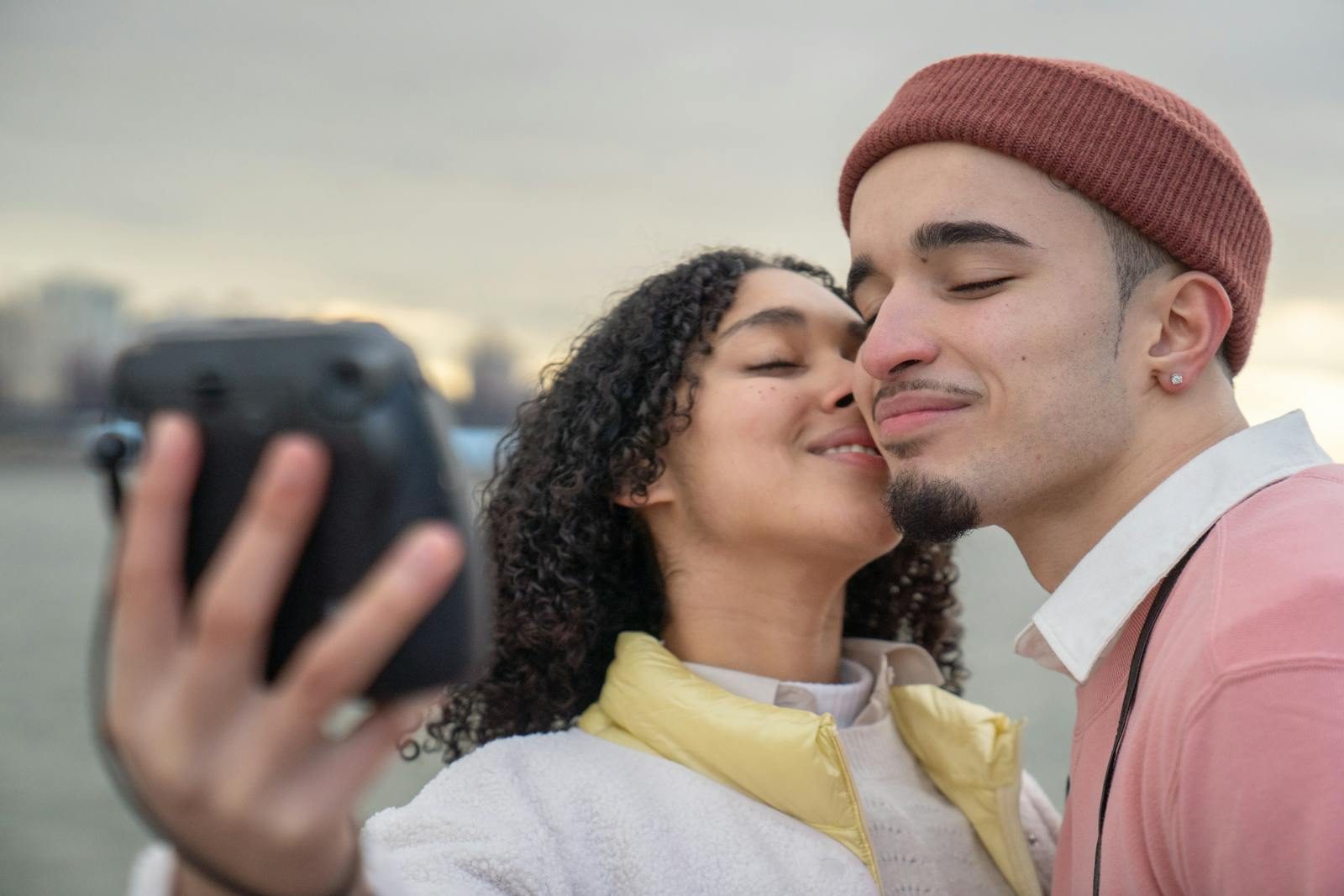 Cheerful Hispanic couple with closed eyes taking self portrait while standing on embankment on blurred background during romantic date in city