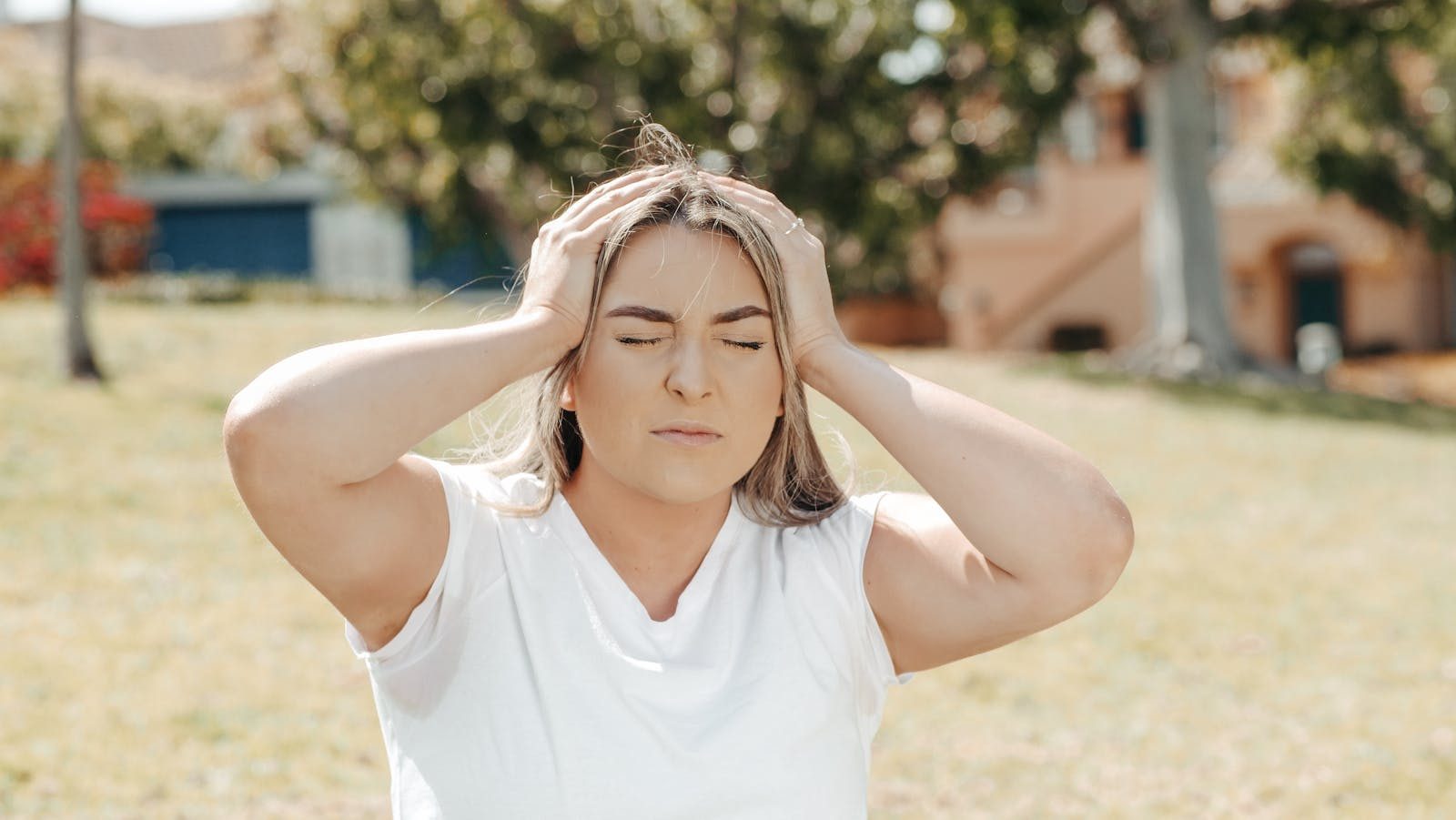 Adult woman holding her head in pain, sitting outdoors under sunlight, likely experiencing a migraine.