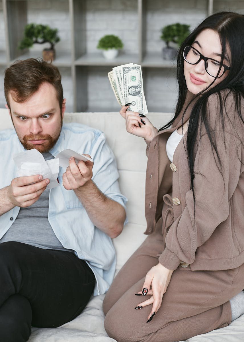 An interracial couple sitting indoors managing finances with cash and receipts, expressing varied emotions.