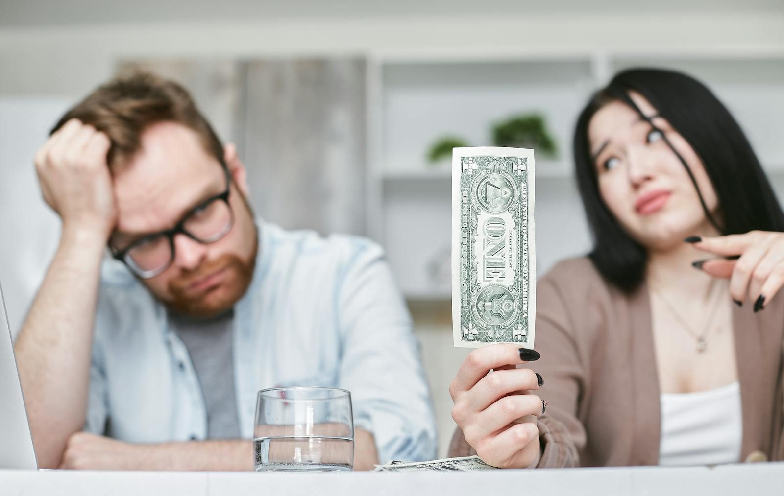 An adult interracial couple expressing concern about finances as they hold a one dollar bill indoors.
