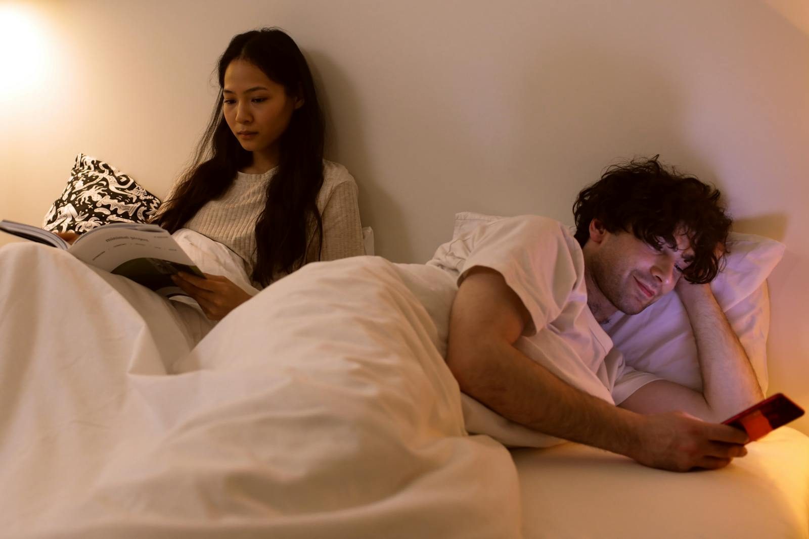 An intimate indoor scene of a couple in bed, reading and using a phone.