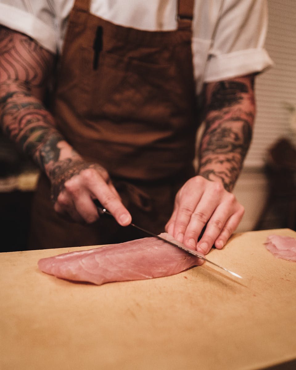 Tattooed chef skillfully slicing raw meat on a wooden cutting board. Indoor setting.