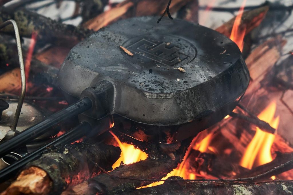 Close-up of a pie iron cooking over a campfire, surrounded by flames and logs.