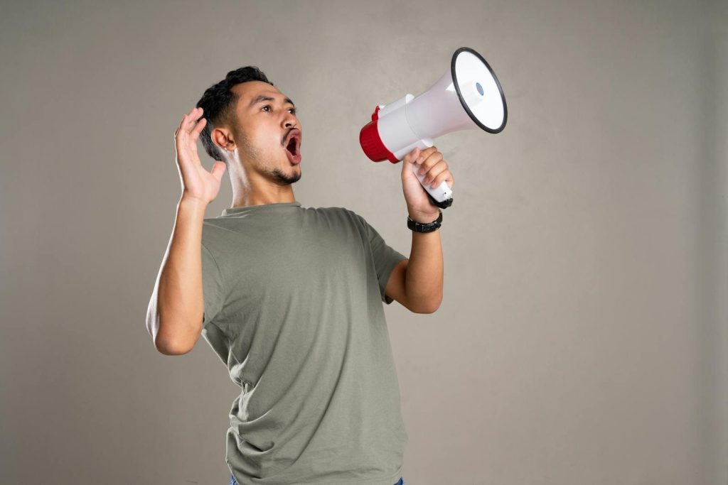 Asian man passionately shouting into a megaphone indoors, conveying energy and excitement.