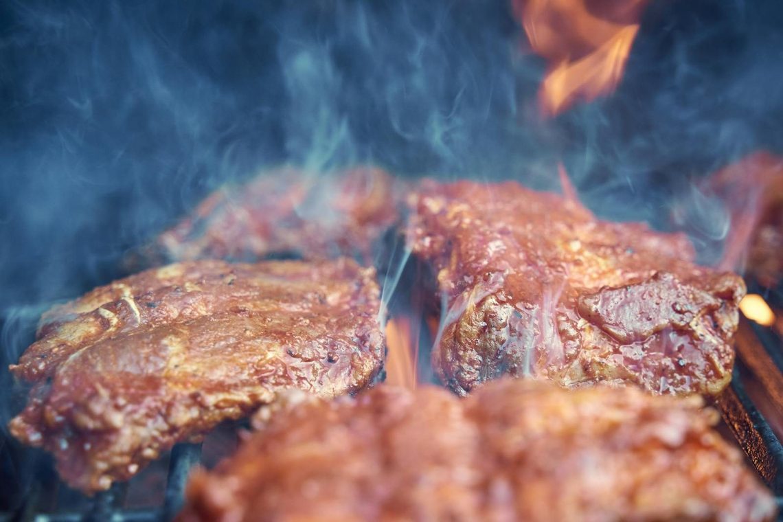 Close-up of sizzling raw meat being grilled with smoke and flames on outdoor barbecue.