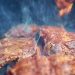 Close-up of sizzling raw meat being grilled with smoke and flames on outdoor barbecue.