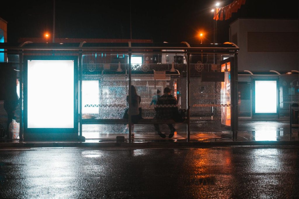 People waiting at an illuminated bus stop on a rainy night, reflecting city lights.
