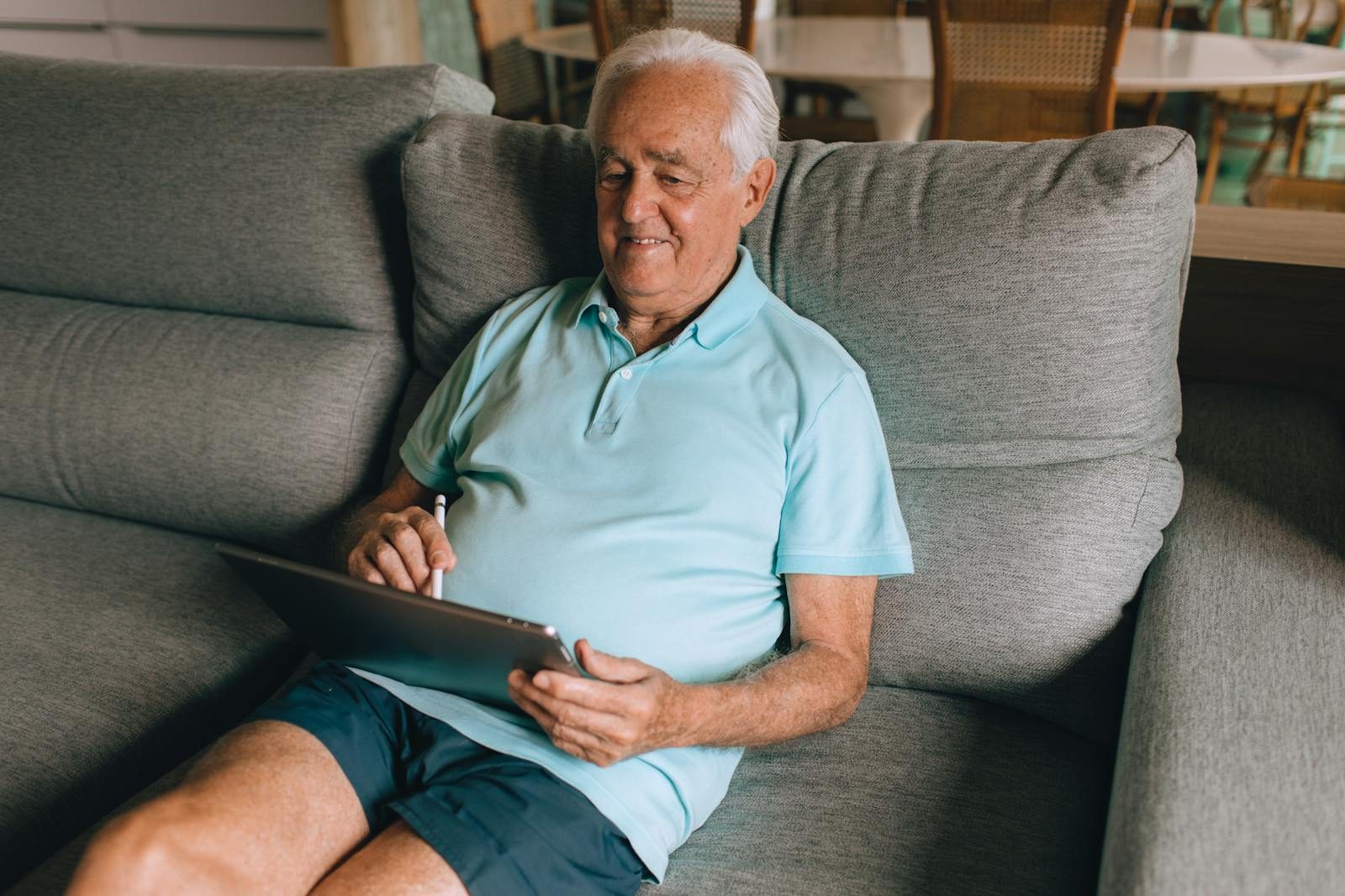 Elderly man enjoying tablet on comfortable sofa indoors, smiling.