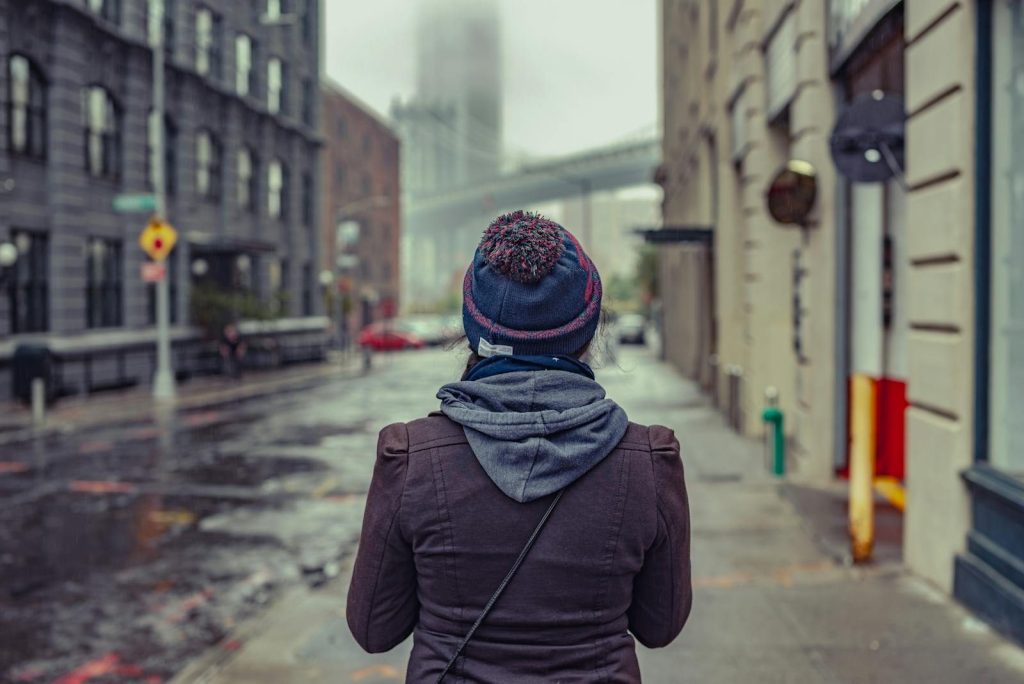 A solitary figure walks through a rainy street in Brooklyn, New York, with the Manhattan Bridge in the background.