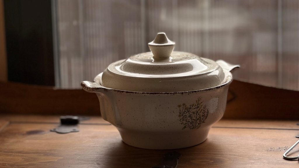 Cozy, rustic ceramic casserole dish placed on a wooden table by a window.