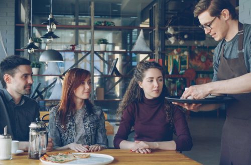 Friendly waiter serving food to young adults in a stylish cafe setting.