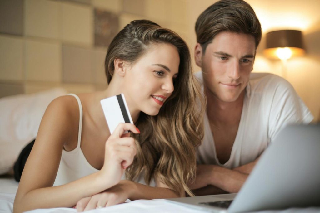 Smiling couple in casual wear lying on bed at home with laptop and making orders in online store with credit card