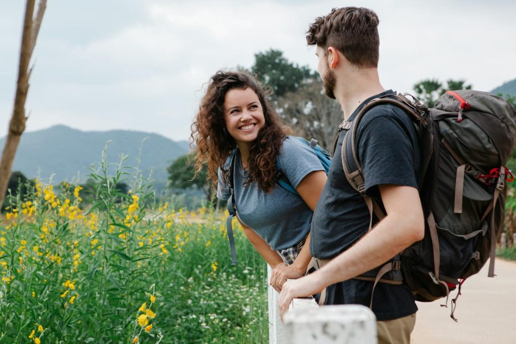 Happy young couple of tourists in casual clothes with backpacks looking at each other while leaning on wooden fence near grassy field with wildflowers in daytime near mountains and trees