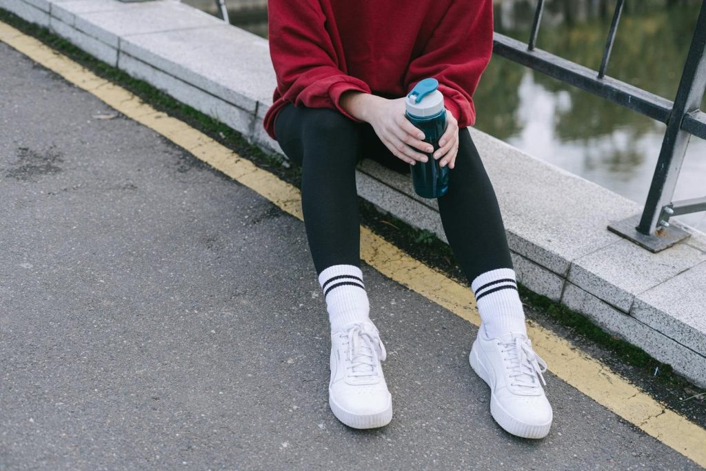 Person sitting on a street curb holding a sports water bottle, wearing sneakers.