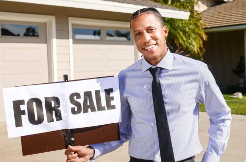 Real estate agent standing with a 'For Sale' sign in front of a house on a sunny day.