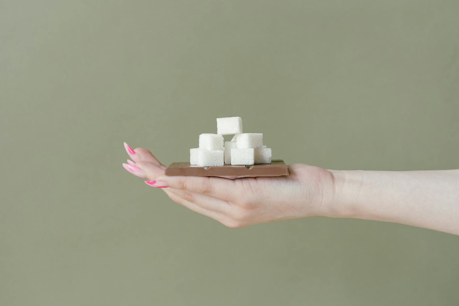 Close-up of a hand holding sugar cubes stacked on a chocolate bar against a plain background.