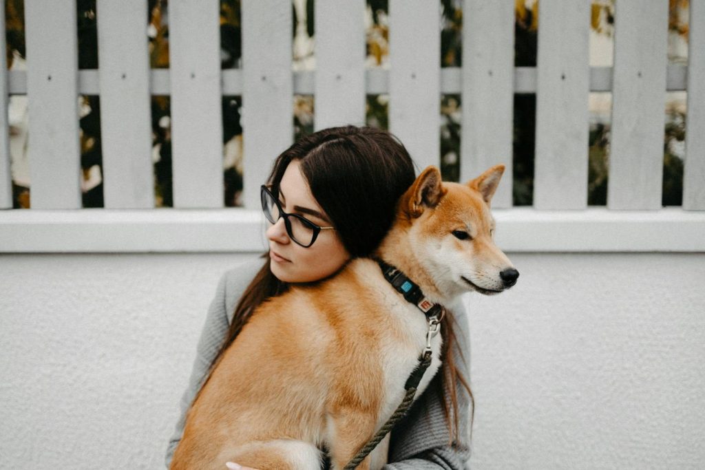 A young woman with glasses cuddles a Shiba Inu dog outdoors by a white fence.